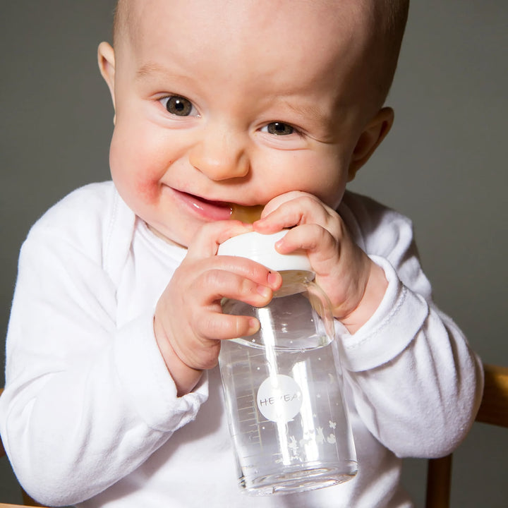 Smiling baby holding a HEVEA glass bottle with a natural rubber nipple, safe for teething, BPA-free, and designed for a natural feeding experience.