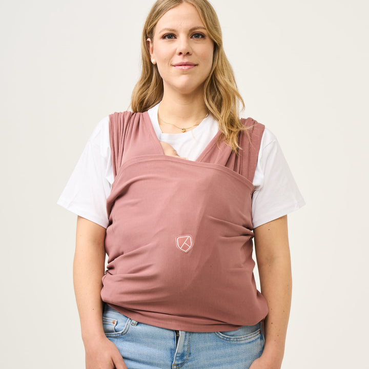 A mother wearing the Koala Babycare Koala Cuddle Band 2 in earth red, carrying her baby comfortably against her chest.