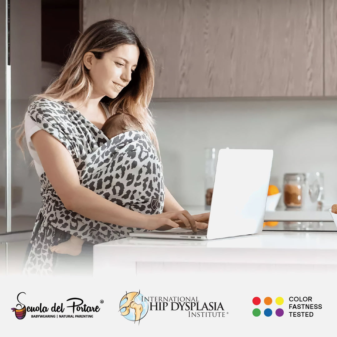 Mom using laptop while baby sleeps in Koala Cuddle Band 2 leopard print baby carrier in kitchen setting.