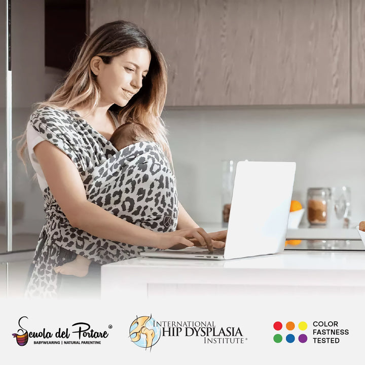 Mom using laptop while baby sleeps in Koala Cuddle Band 2 leopard print baby carrier in kitchen setting.