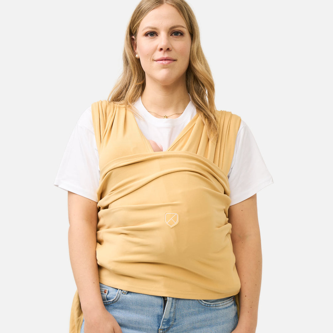 Koala Babycare Koala Cuddle Band 2 baby carrier in mustard yellow, shown worn by woman with infant