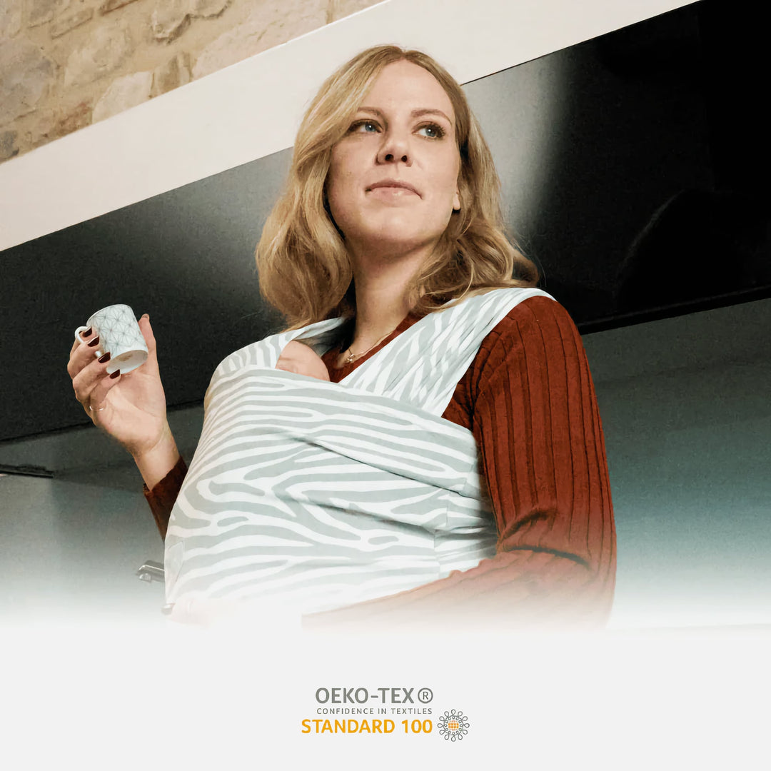 Mother wearing the Koala Cuddle Band 2 baby sling carrier in tiger-print gray, holding a coffee cup while keeping her baby close. OEKO-TEX Standard 100 certified for safe and toxin-free fabric.