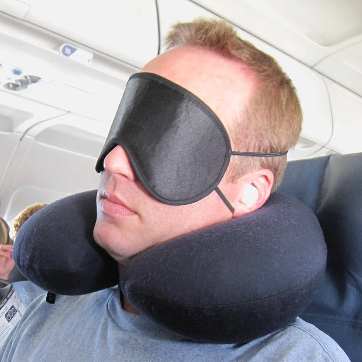 Man wearing Mack's Shut-Eye Shade Sleep Mask and earplugs on an airplane with a neck pillow for travel rest.
