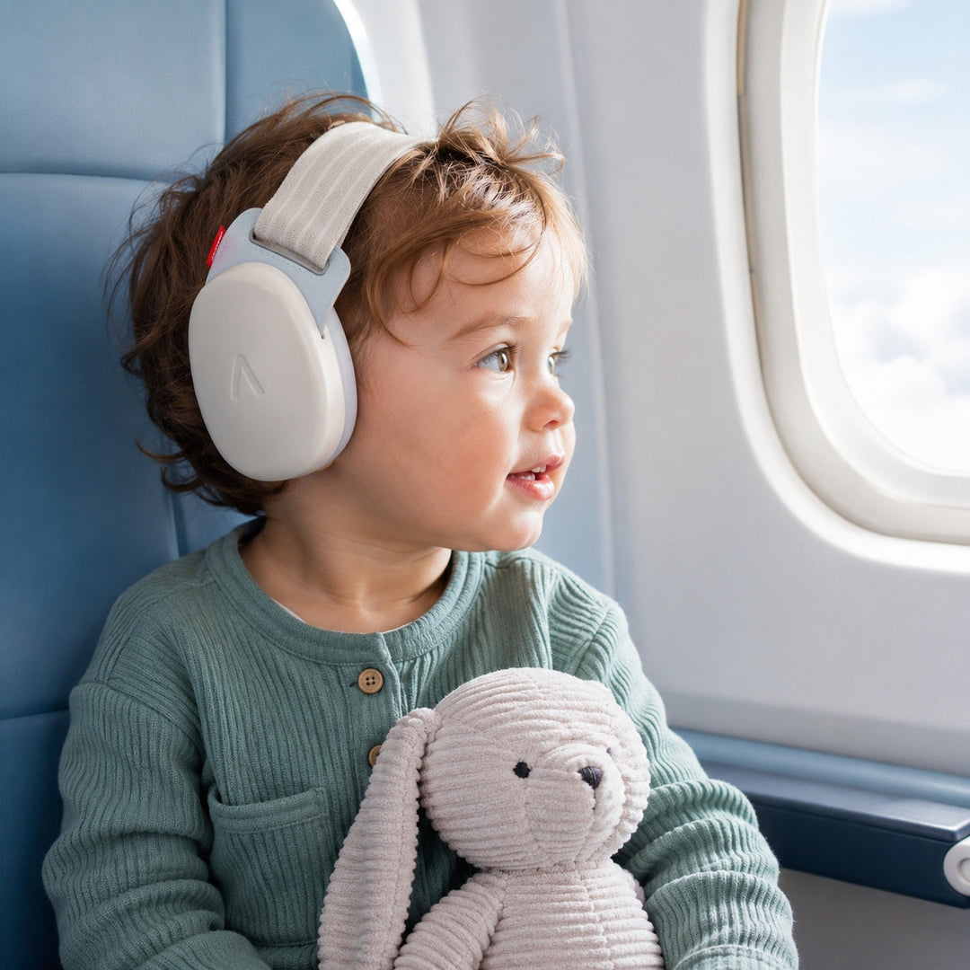 Toddler wearing sand Alpine baby earmuffs by an airplane window while holding a plush toy