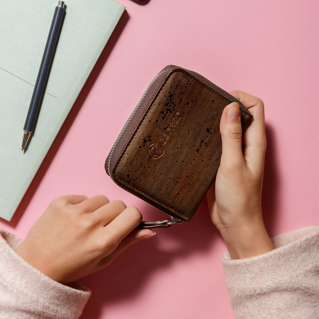 Hands pulling zipper on Corkor dark brown cork square wallet on pink desk with notebook and pen