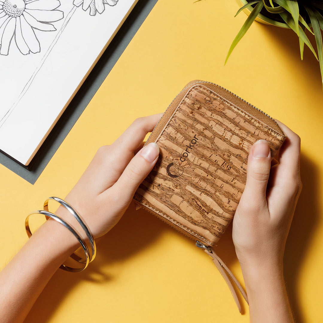 Hands holding Corkor Zebra cork square wallet on a yellow desk beside floral artwork and a plant