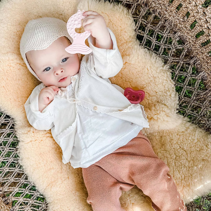 Baby lying on soft mat holding HEVEA Fish Teether in Powder Pink, natural rubber toy for soothing teething discomfort