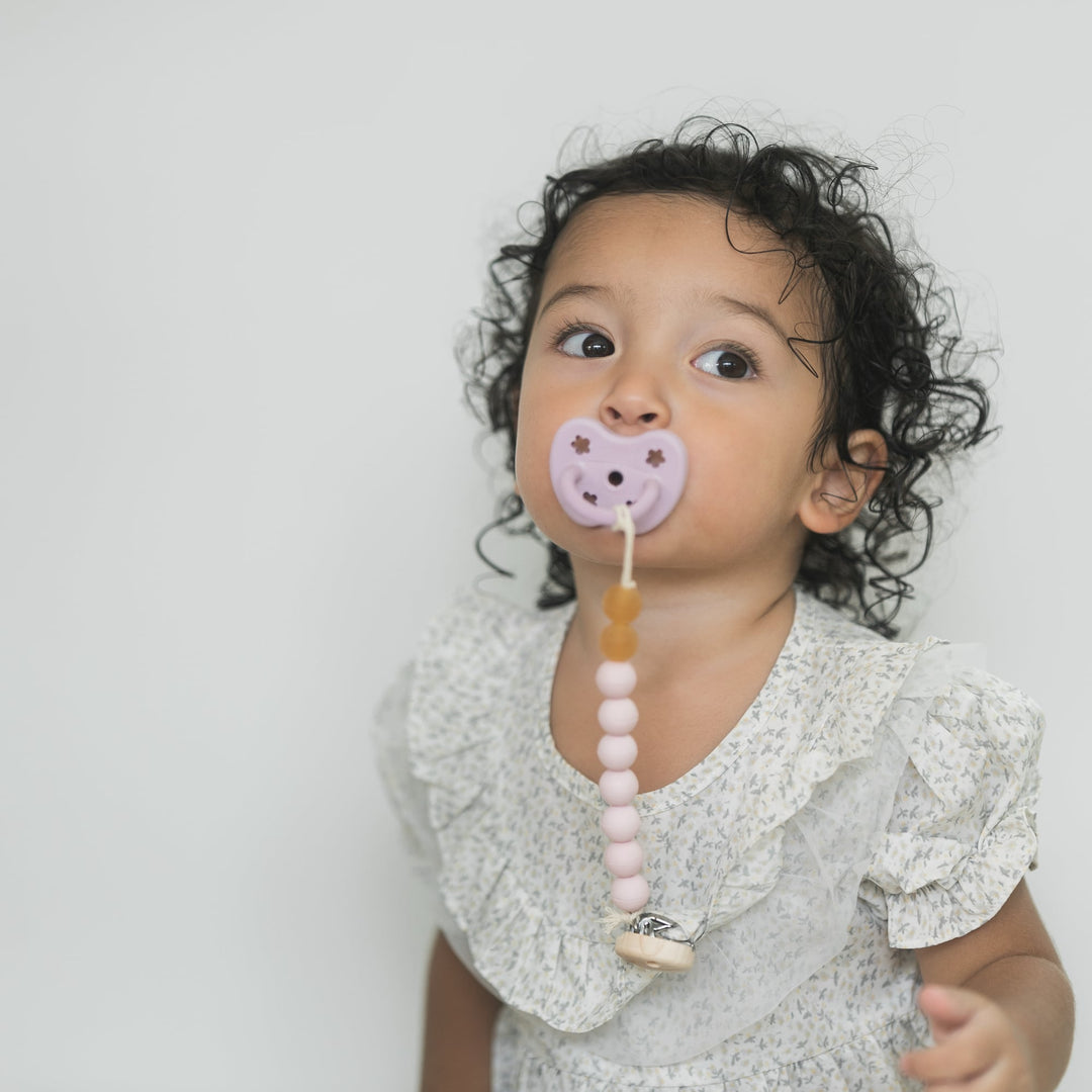 Child wearing a Hevea Powder Pink pacifier holder with pink and amber beads, wooden clip and attached pacifier