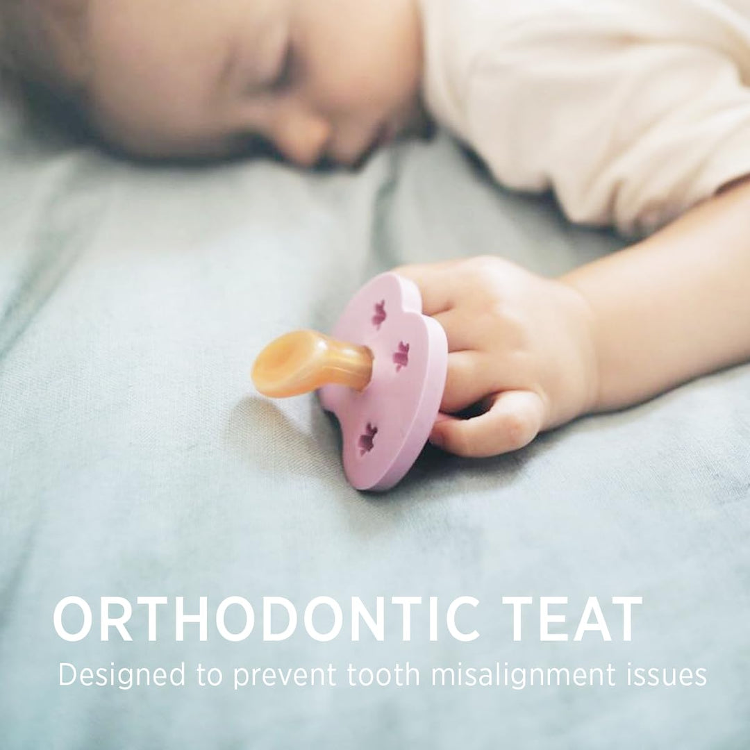 Baby holding pink Hevea orthodontic pacifier with natural rubber teat in a soft sleep setting