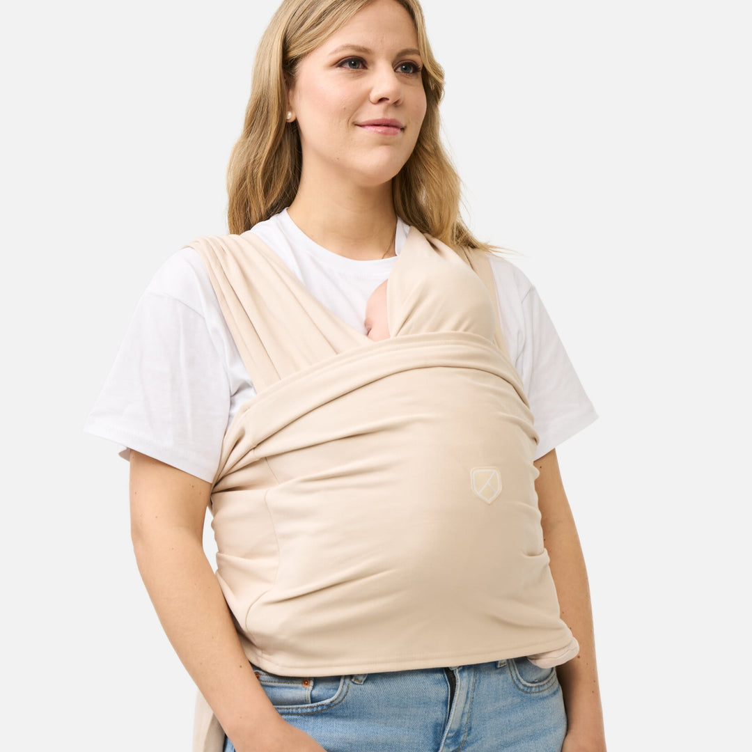 Side view of a mother wearing the Koala Cuddle Band 2 baby sling carrier in beige, keeping her baby secure and close in an ergonomic position.