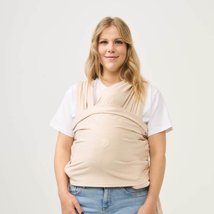 Mother wearing the Koala Cuddle Band 2 baby wrap carrier in beige, holding her baby securely in a comfortable and ergonomic position.