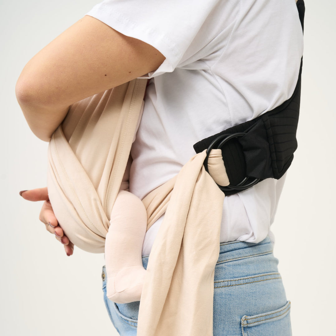 Side view of a mother adjusting the Koala Cuddle Band 2 baby carrier in beige, showing the ergonomic support and secure ring system.