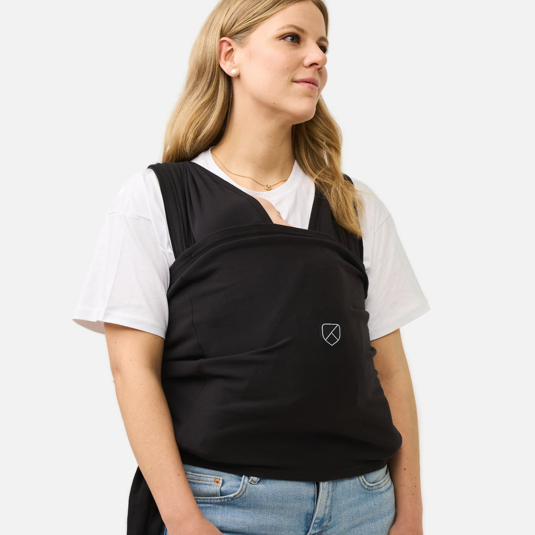 Mother wearing the Koala Cuddle Band 2 baby wrap carrier in black, keeping her baby secure and comfortable in an ergonomic position.