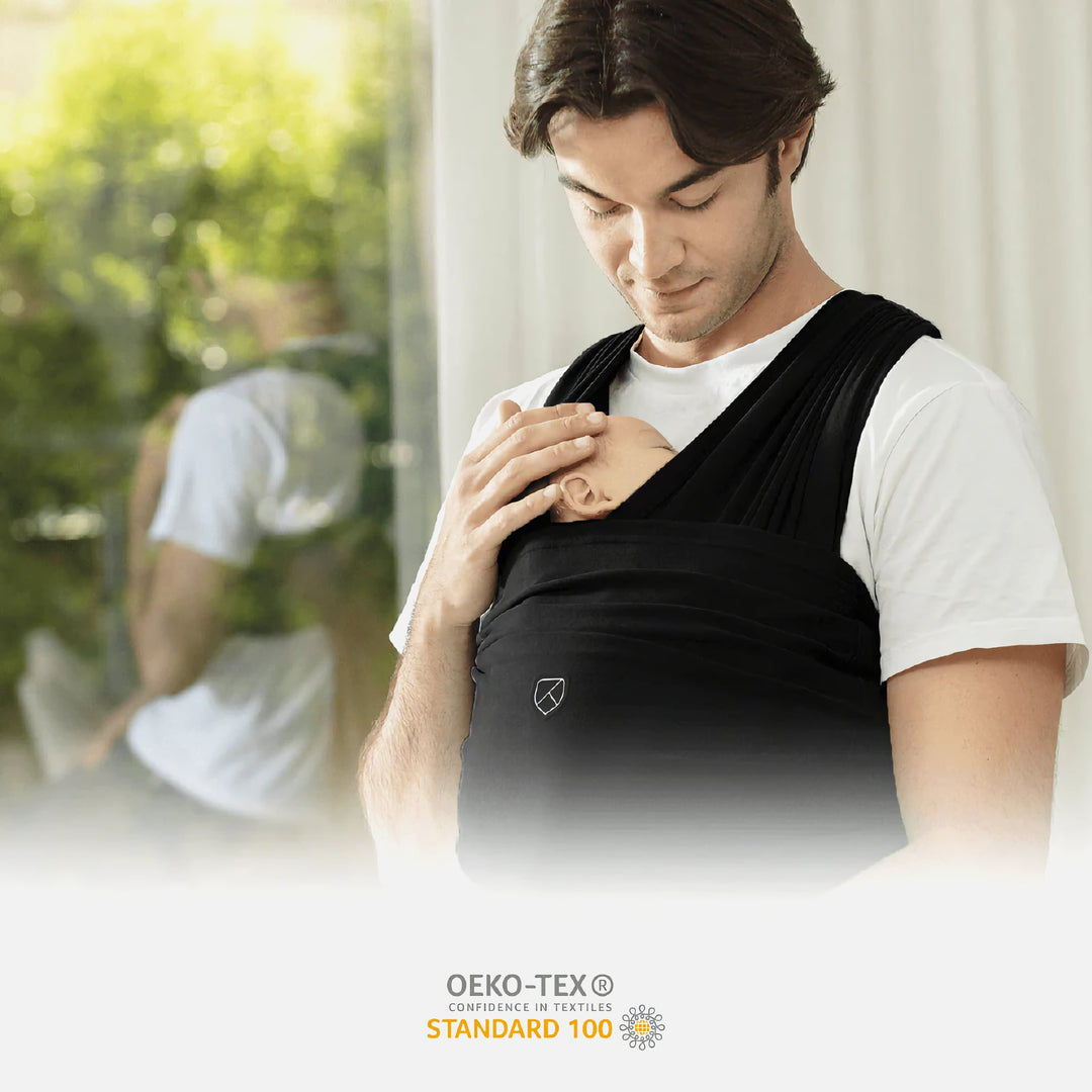 Father wearing the Koala Cuddle Band 2 baby wrap in black, holding a newborn close to his chest. OEKO-TEX® Standard 100 certified for safety and comfort.