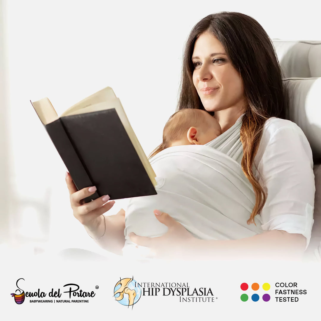 Mother wearing the Koala Cuddle Band 2 baby sling while reading, with certification logos for babywearing safety, hip health, and color fastness.