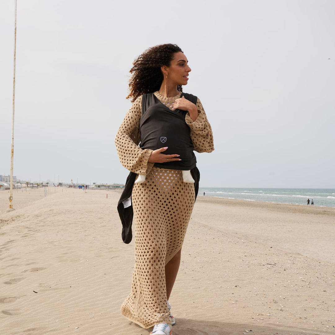 Mother walking on the beach wearing Koala Babycare Koala Cuddle Band 2 in military green, carrying her baby comfortably.