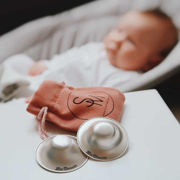 Silver Mamas 925 sterling silver nursing cups with a pink storage bag, placed near a baby in the background.